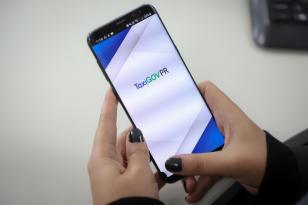 Pessoa com unhas pintadas com cor escura manuseia celular; tela do TaxiGOVPR está aberta