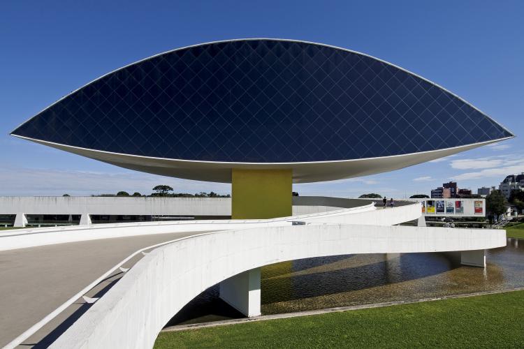 Imagem do Museu Oscar Niemeyer, popularmente conhecido como "Museu do Olho" devido ao seu formato icônico, destaca a arquitetura contemporânea que caracteriza Curitiba. Projetado pelo renomado arquiteto Oscar Niemeyer, o museu foi idealizado durante o último mandato de Jaime Lerner como governador do Paraná (1999-2002) e inaugurado em 22 de novembro de 2002.