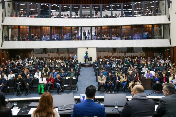 Em primeiro plano, mesa de abertura de costas para a foto. Em segundo plano, a plateia presta atenção.