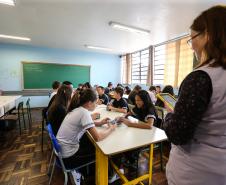 Professora em pé dando aula para alunos sentados, dentro de sala de aula