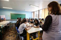 Professora em pé dando aula para alunos sentados, dentro de sala de aula