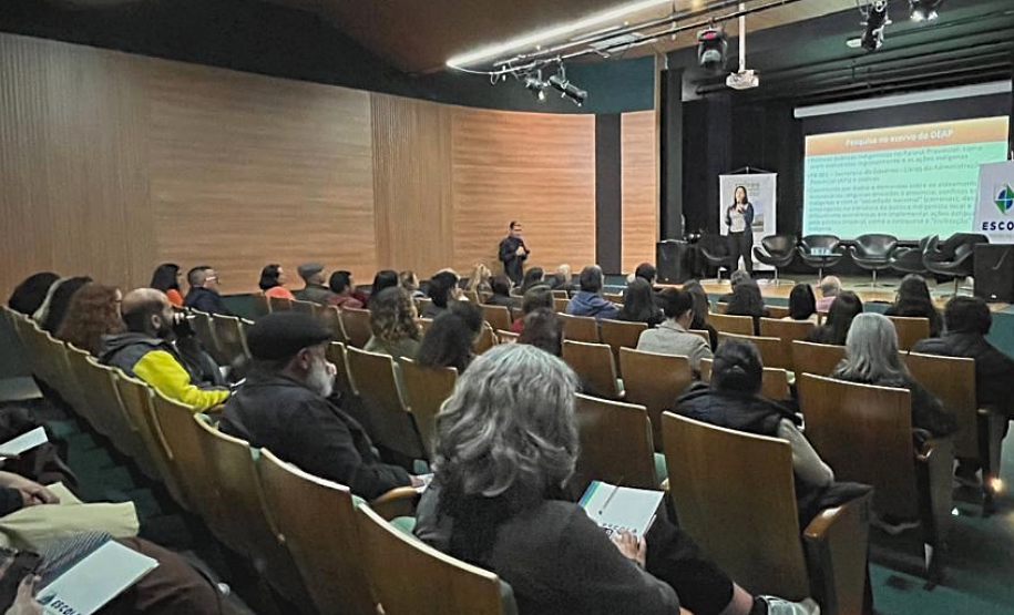 Público sentado em auditório assiste palestrante Marina Braga, que está em cima de um palco.