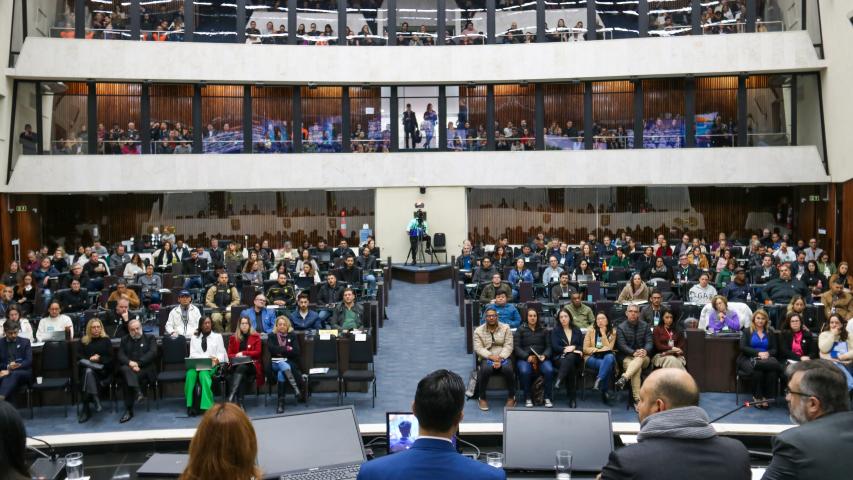 Em primeiro plano, mesa de abertura de costas para a foto. Em segundo plano, a plateia presta atenção.