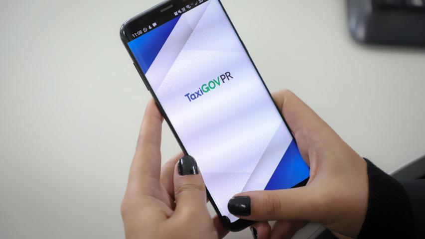 Pessoa com unhas pintadas com cor escura manuseia celular; tela do TaxiGOVPR está aberta